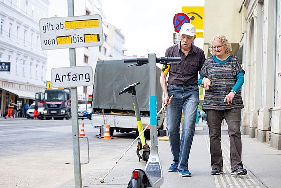 älterer Mann mit Langstock eingehakt bei einer Frau geht auf E-Scooter mitten am Gehsteig zu