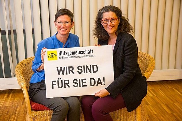 Zwei Mitarbeiterinnen der Hilfsgemeinschaft sitzen auf je einem Stuhl und halten gemeinsam lächelnd ein Schild mit den Worten "Wir sind für Sie da!" in die Kamera.