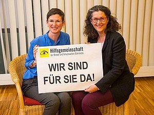 Zwei Mitarbeiterinnen der Hilfsgemeinschaft sitzen auf je einem Stuhl und halten gemeinsam lächelnd ein Schild mit den Worten "Wir sind für Sie da!" in die Kamera.