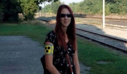 Frau mit Armschleife, Sonnenbrille und Taststock steht und lächelt in Kamera