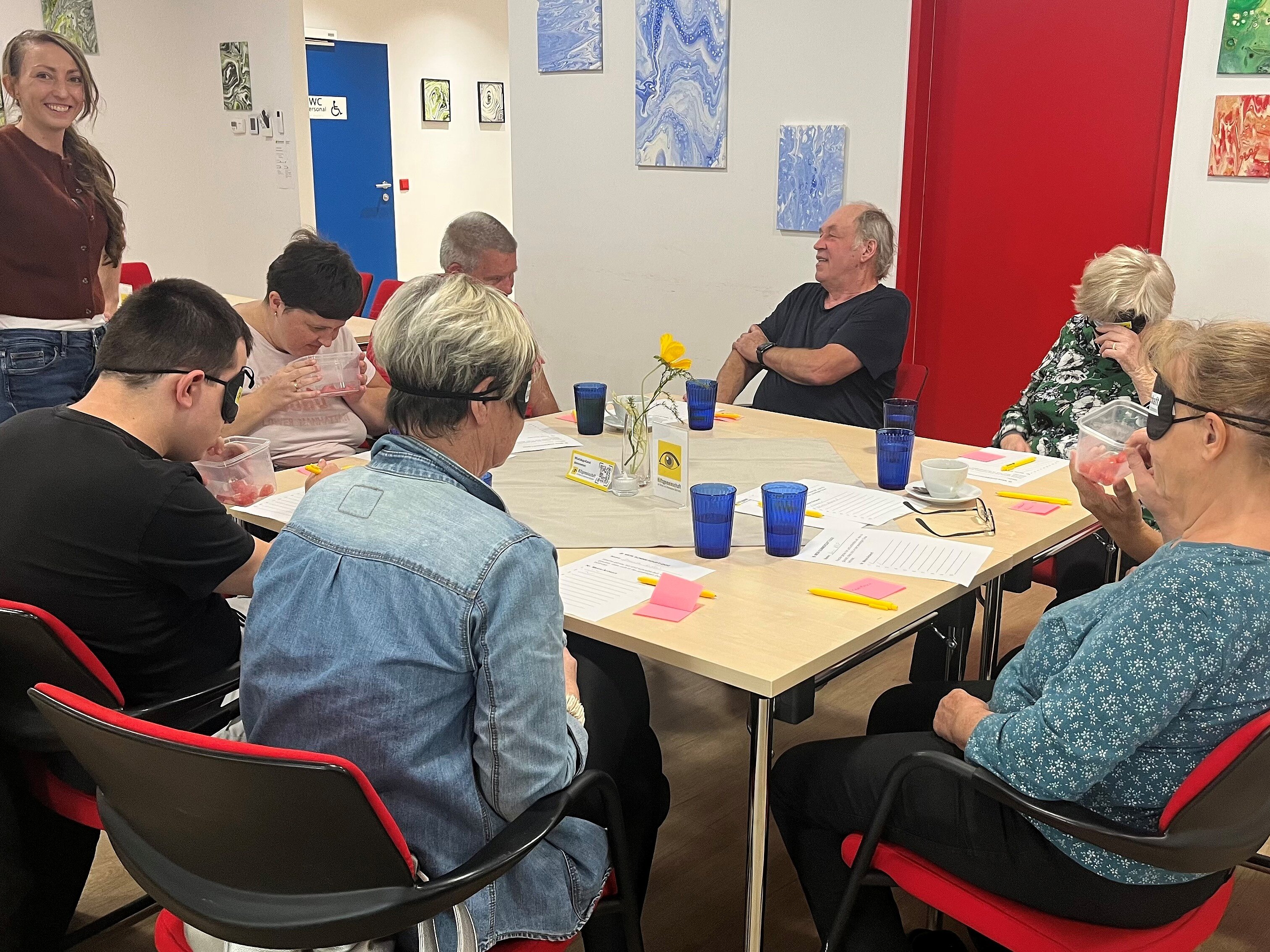 Foto: © HG / Hlava Eine Gruppe von Menschen sitzt bei einem Workshop an einem Tisch. Einige tragen Simulationsbrillen und bearbeiten gemeinsam Unterlagen; eine Frau steht daneben. Der Raum ist mit abstrakten Bildern, roten Türen und blauen Gläsern ausgestattet.