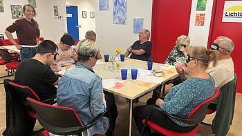 Eine Gruppe von Menschen sitzt bei einem Workshop an einem Tisch. Einige tragen Simulationsbrillen und bearbeiten gemeinsam Unterlagen; eine Frau steht daneben. Der Raum ist mit abstrakten Bildern, roten Türen und blauen Gläsern ausgestattet.