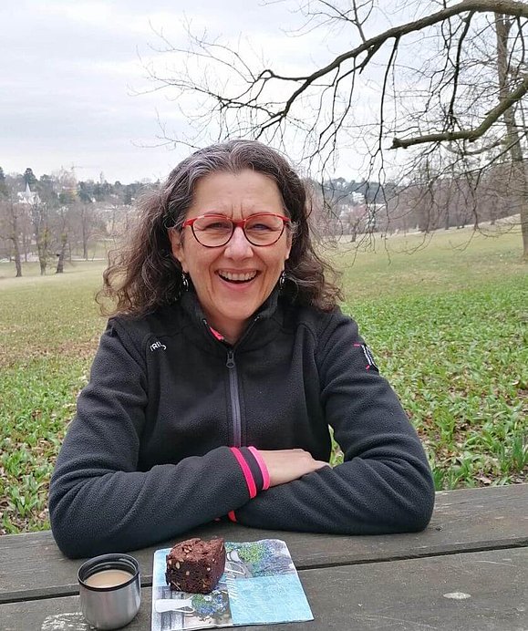 Eine Frau mit dunklen gelockten Haaren und roter Brille sitzt auf einer Parkbank, vor sich ein Stück Kuchen und Kaffee. 