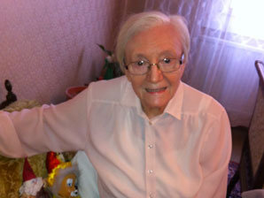 Eine ältere Dame sitzt fröhlich lächelnd auf einem Sofa, neben ihr bunte Puppen