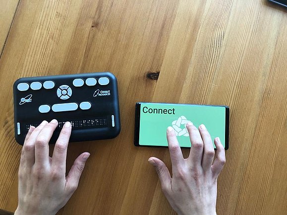 Hände bedienen eine Braillezeile und die Oberfläche eines Smartphones