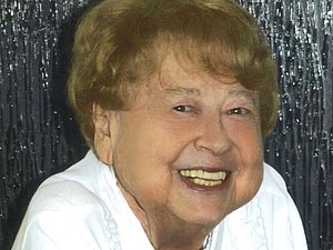 ältere Frau mit weißem Shirt und dunkelblonden Haaren lächelt in die Kamera, Hintergrund in silber