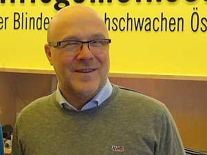 Daniele Marano in einem grauen Pullover und blauem Hemd, im Hintergrund eine gelbe Wand mit schwarzer Schrift.