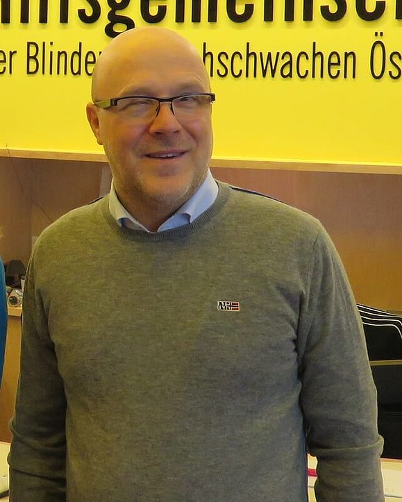 Daniele Marano in einem grauen Pullover und blauem Hemd, im Hintergrund eine gelbe Wand mit schwarzer Schrift.