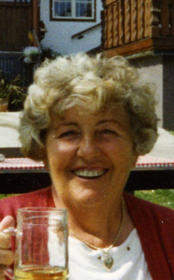 Eine ältere Dame mit Dauerwelle hält ein Glas in der Hand und lächelt fröhlich in die Kamera, hinter ihr sieht man einen Teil eines alten Bauernhauses