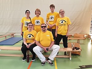 Sechs Personen mit gelben T-Shirts der Hilfsgemeinschaft stehen in einem Turnsaal für ein Foto zusammen. 