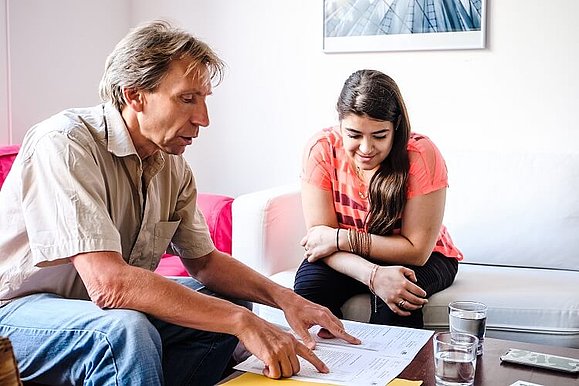 Ein Mann und eine junge Frau sitzen bei einem Couchtisch, vor ihnen Dokumente die sie gemeinsam durchgehen. Der Mann zeigt mit seinen Zeigefingern auf den Text. 