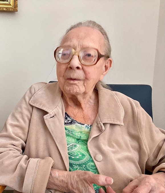Ältere Frau mit Brille und beiger Jacke sitzt auf Stuhl