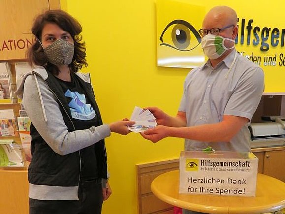 Eine Frau und ein Mann stehen sich gegenüber. Beide tragen eine Maske. Die Frau gibt dem Mann einige REWE Einkaufsgutscheine.