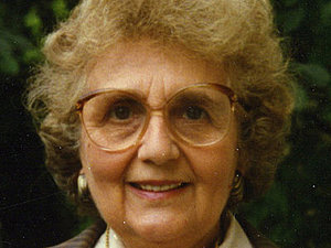 Eine ältere Dame mit großer Brille steht vor einer grünen Hecke und lächelt freundlich in die Kamera 