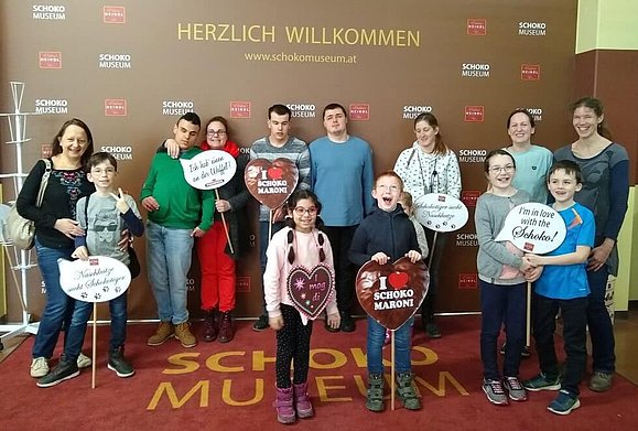 14 Personen stehen nebeneinander vor der Willkommenswand des Schokomuseum. Einige Kinder halten Schilder mit Sprüchen in der Hand.