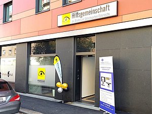 Eingangstür von außen mit gelben Luftballons, Hilfsgemeinschaft-Schild und Aufsteller mit Videbis Logo