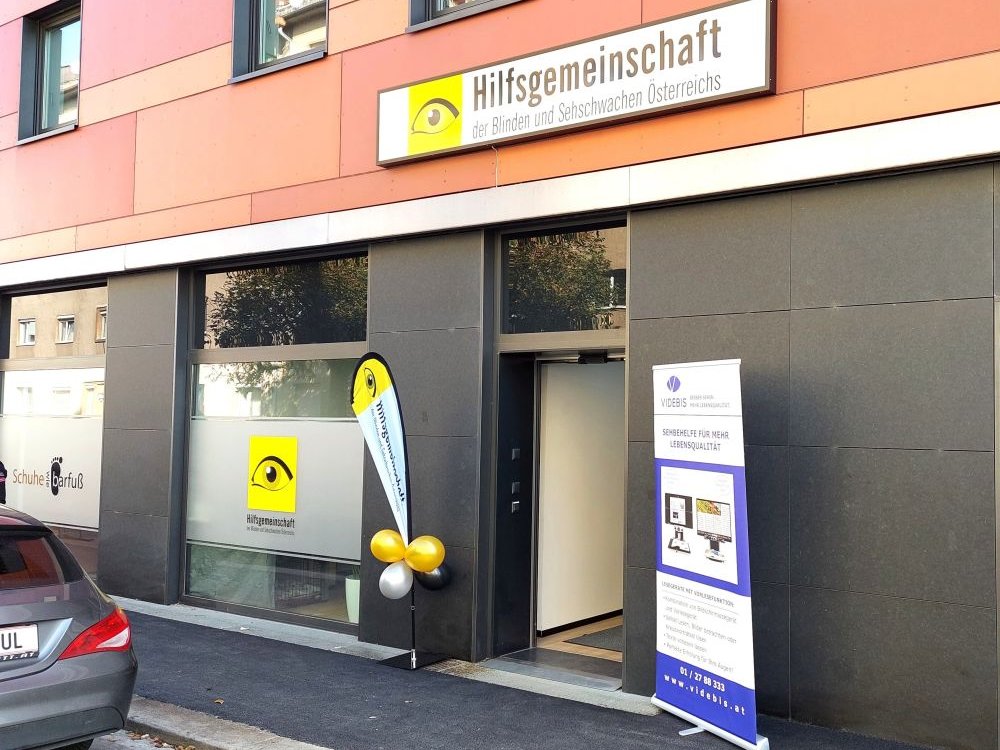 Eingangstür von außen mit gelben Luftballons, Hilfsgemeinschaft-Schild und Aufsteller mit Videbis Logo