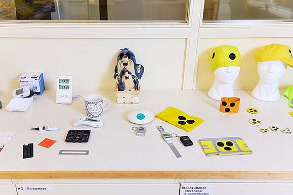 Auf einem Tresen liegen viele unterschiedliche Hilfsmittel wie kennzeichnende Armbinden, Buttons und sogar Badehauben, sprechende elektronische Geräte und vieles mehr.