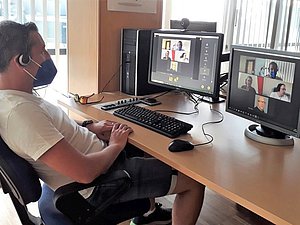Mann betrachtet zwei Computerbildschirme, auf denen ein Multi-User-Videocall geöffnet ist.