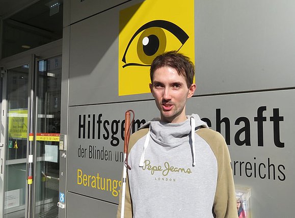 David Erkinger muss sich an seiner Uni mit vielen Barrieren wie nicht-barrierefreien Dokumenten herumschlagen. Dabei unterstützt ihn die Hilfsgemeinschaft. junger Mann mit kurzen braunen Haaren vor Hauseingang mit gelbem Logo Auge im Hintergrund