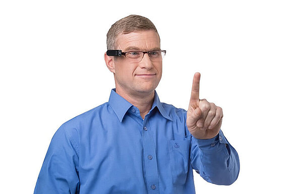 Mann in blauem Hemd hält seinen Zeigefinger in die Luft, auf seinem Kopf eine Brille mit OrCam, Copyright: OrCam VIDEBIS