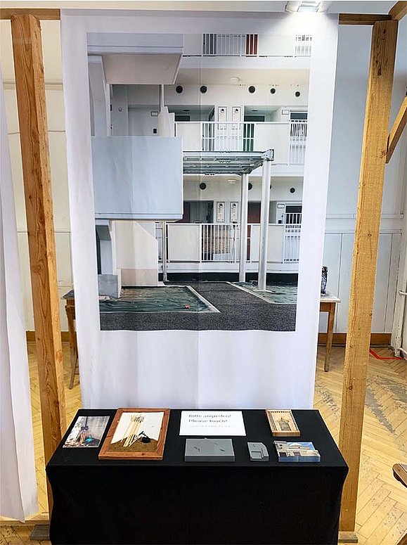 Foto eines Ausstellungsdisplays: auf einem hohen Holzrahmen hängt groß ein Architekturfoto auf Papier. Es zeigt ein weißes Gebäude mit Balkonen. Davor steht ein Tisch mit schwarzem Überwurf, auf dem taktile Fotografien liegen. 
