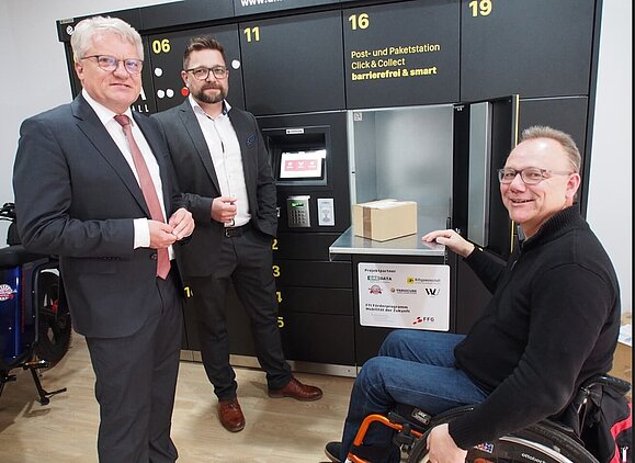 Rollstuhlfahrer mit zwei elegant gekleideten Herren vor Paketbox