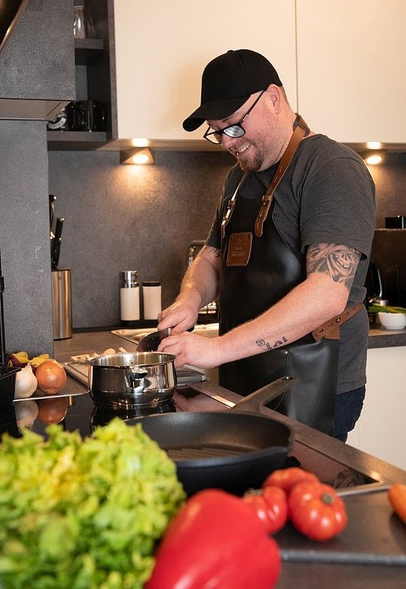 Mann mit schwarzem Cap und Kochschürze schneidet Gemüse in der Küche