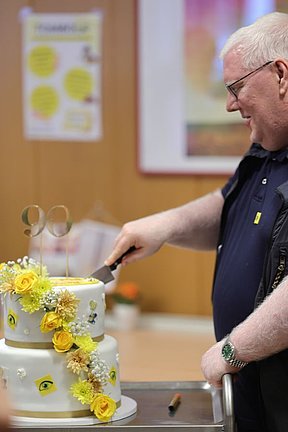 90 Jahre Hilfsgemeinschaft – das muss gefeiert werden Ein Mann (Elmar Fürst) schneidet eine Torte an, auf welcher die Zahl 90 ist.