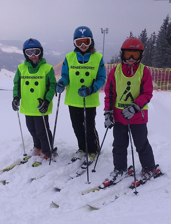 drei Kinder auf Skiern mit Helm und Skibrille sowie neongelbem Trikot mit 3 Punkten und Aufschrift SEHBEHINDERT