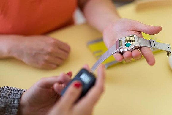 Nahaufnahme der Hände zweier Personen, die eine spezielle Uhr betrachten. Eine Person hält die Uhr, während die andere ein kleines elektronisches Gerät hält. Die Uhr hat ein metallisches Armband und ein digitales Display. 