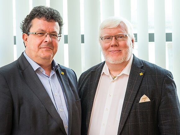 Zwei Männer im dunklen Anzug stehen nebeneinander.