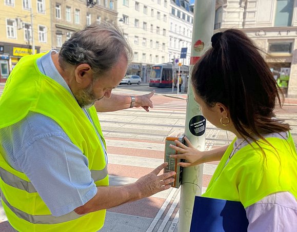 Mann erklärt junger Frau die Funktion der Akustikampel und zeigt auf den Straßenübergang