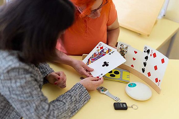 Zwei Frauen betrachten Spielkarten in Riesengröße. Sie halten gemeinsam einige der Karten in der Hand, die anderen Karten stecken in einem Ständer, der vor ihnen auf dem Tisch steht. Auf dem Tisch liegen weitere Hilfsmittel. 