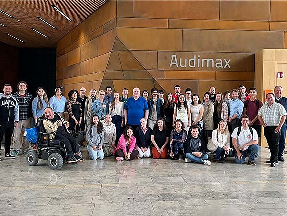 Eine große Gruppe von Teilnehmerinnen und Teilnehmern der Summer School 2025 steht und sitzt gemeinsam für ein Gruppenfoto im Foyer eines Gebäudes mit der Beschriftung "Audimax". Im Vordergrund ist eine Person im Elektrorollstuhl. Alle blicken lächelnd in die Kamera.