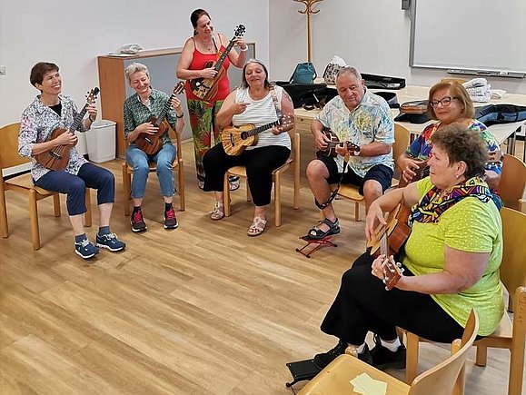 Foto der Ukulele-Gruppe der Hilfsgemeinschaft, sieben Menschen sitzen auf Sesseln und üben Ukulele.