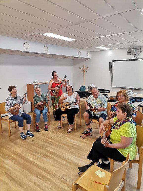 Foto der Ukulele-Gruppe der Hilfsgemeinschaft, sieben Menschen sitzen auf Sesseln und üben Ukulele.