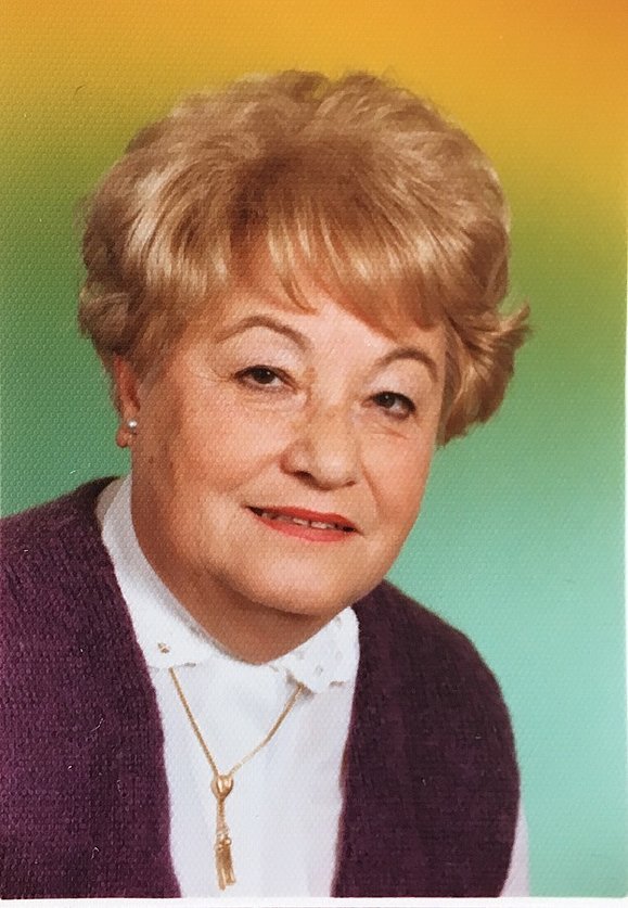 Eine Dame mit blonder Dauerwelle sitzt vor einem bunten Fotohintergrund und lächelt in die Kamera