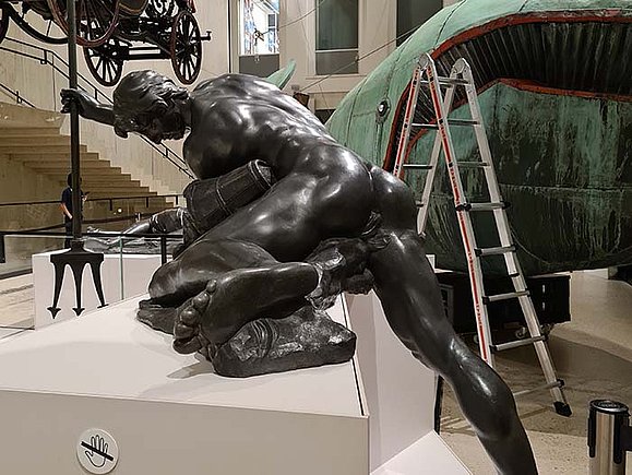 Bronzestatue eines nackten Mannes der vornübergebeugt ist.