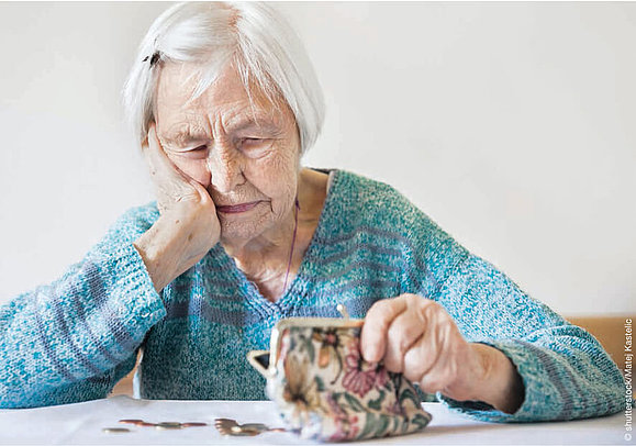 Eine ältere Dame sitzt beim Tisch und schaut verzweifelt in Ihre leere Geldbörse.