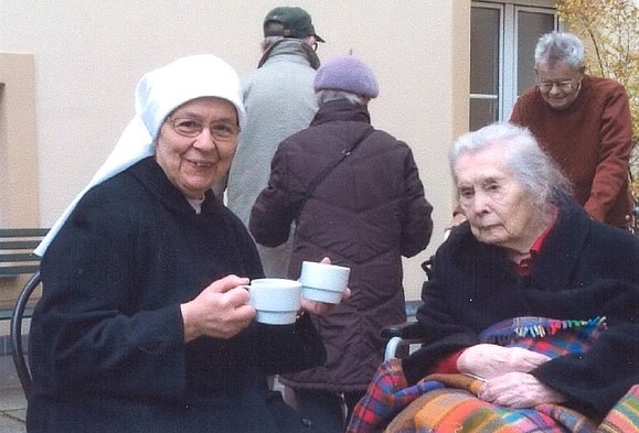 Nonne mit Teetasse in der Hand und ältere Dame mit Decke schauen in die Kamera