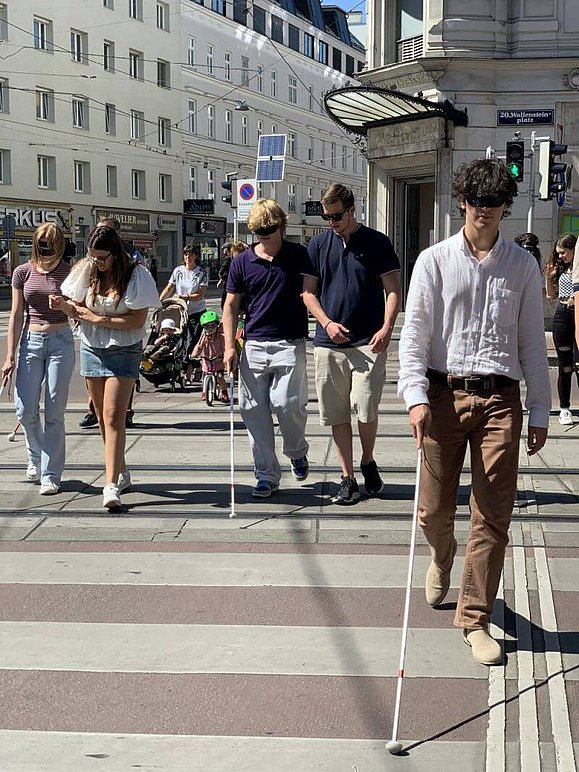 mehrere Junge Menschen, teils eingehakt mit weißem Stock und Augenbinde gehen über Zebrastreifen