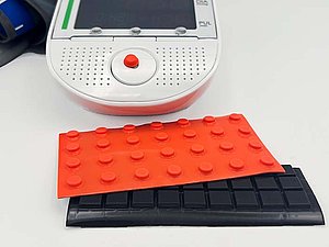 Jeweils ein Bogen mit erhabenen orangen und schwarzen Klebepunkten, dahinter ein Gerät, auf dessen Bedienfeld ein orangener Punkt klebt. 