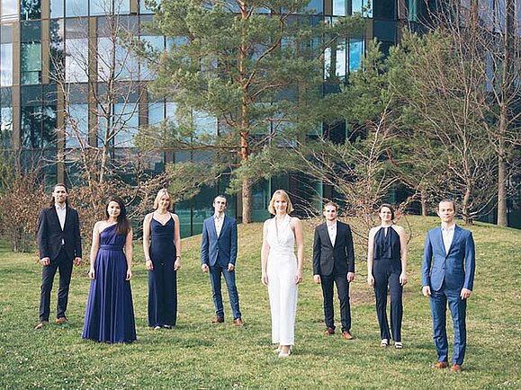 Gruppenfoto des Vocalodie Vokalensembles in blau, schwarz und weißen Outfits auf einem Rasen, im Hintergrund ein Gebäude.