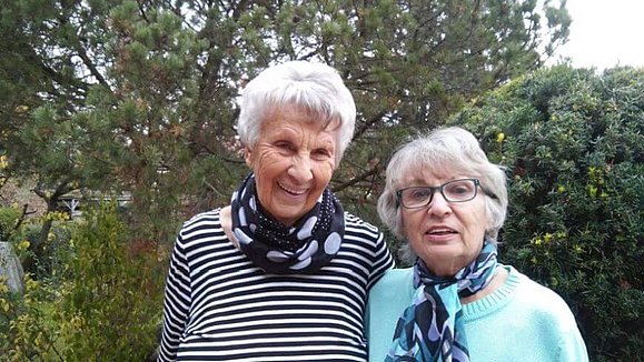 Zwei Seniorinnen stehen eng nebeneinander im Garten und lächeln in die Kamera.