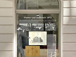 Schaufenster mit Aufdruck: JMW Vision und Aufbruch 1873 - Das israelitische Blindeninstitut auf der Hohen Warte. Unterhalb mehrere schwarz-weiß-Fotos von Räumlichkeiten und eine Zeichnung eines Hauses. am linken Rand: Foto von Mann im Anzug schwarzweiß von hinten