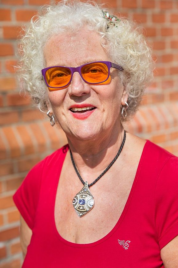 ältere Frau mit weißen Locken, Brillen mit orangefarbigen Gläsern und rotem T-Shirt lächelt in die Kamera