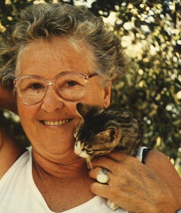 Eine ältere Dame mit Dauerwelle und Brille lächelt fröhlich in die Kamera während sie ein Katzenjunges streichelt, das auf ihrer Schulter sitzt und sich an ihr Gesicht schmiegt