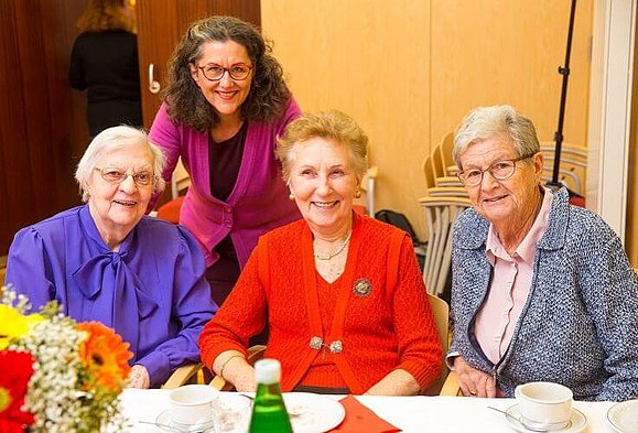 Eine Gruppe von 4 Damen sitzt bei Tisch und lächelt in die Kamera