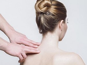 Nackter Rücken einer Frau, zwei Hände deuten auf die Halswirbelsäule.